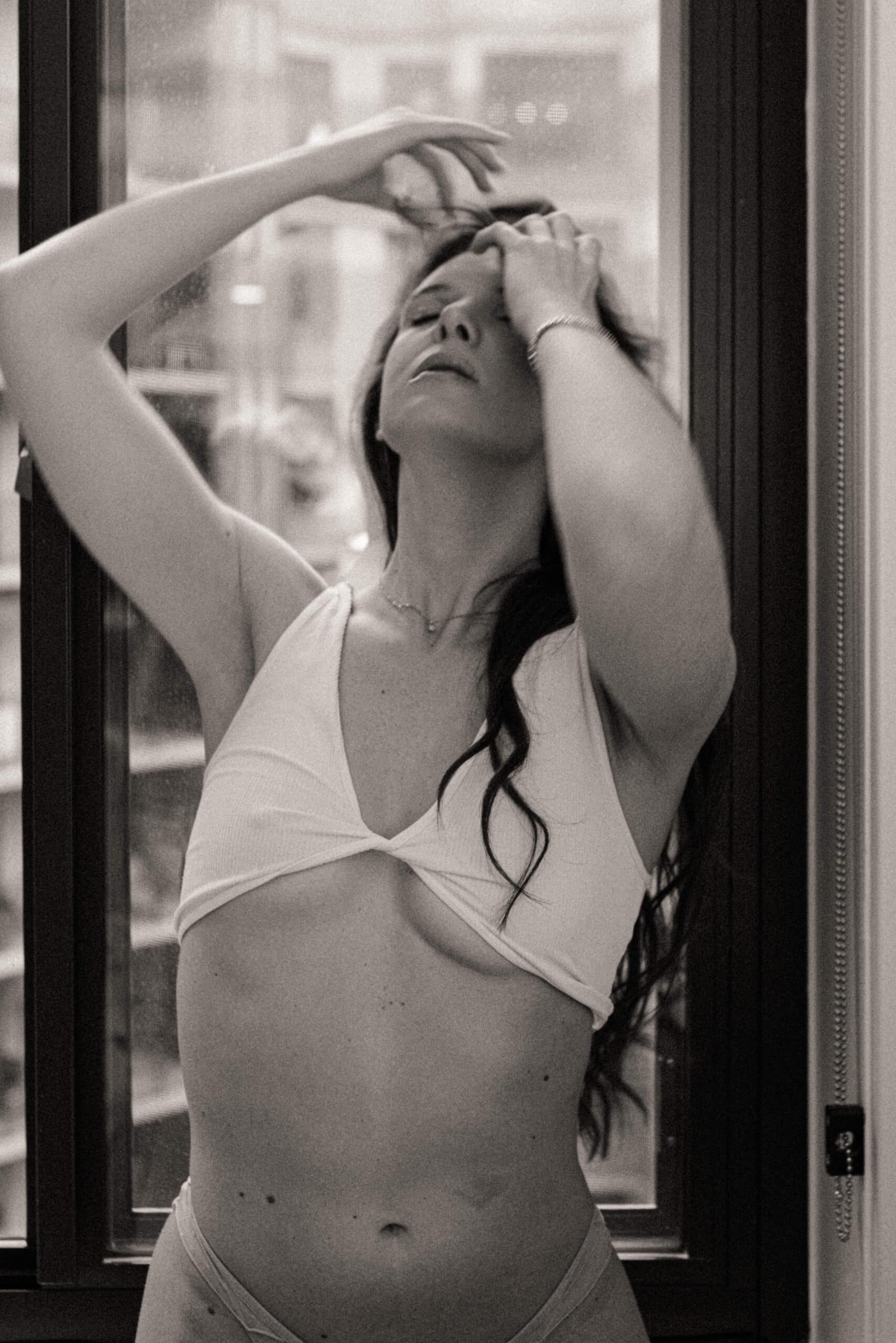 Close-up B&W photo of a woman in underwear with motion-blurred hand in hair by window