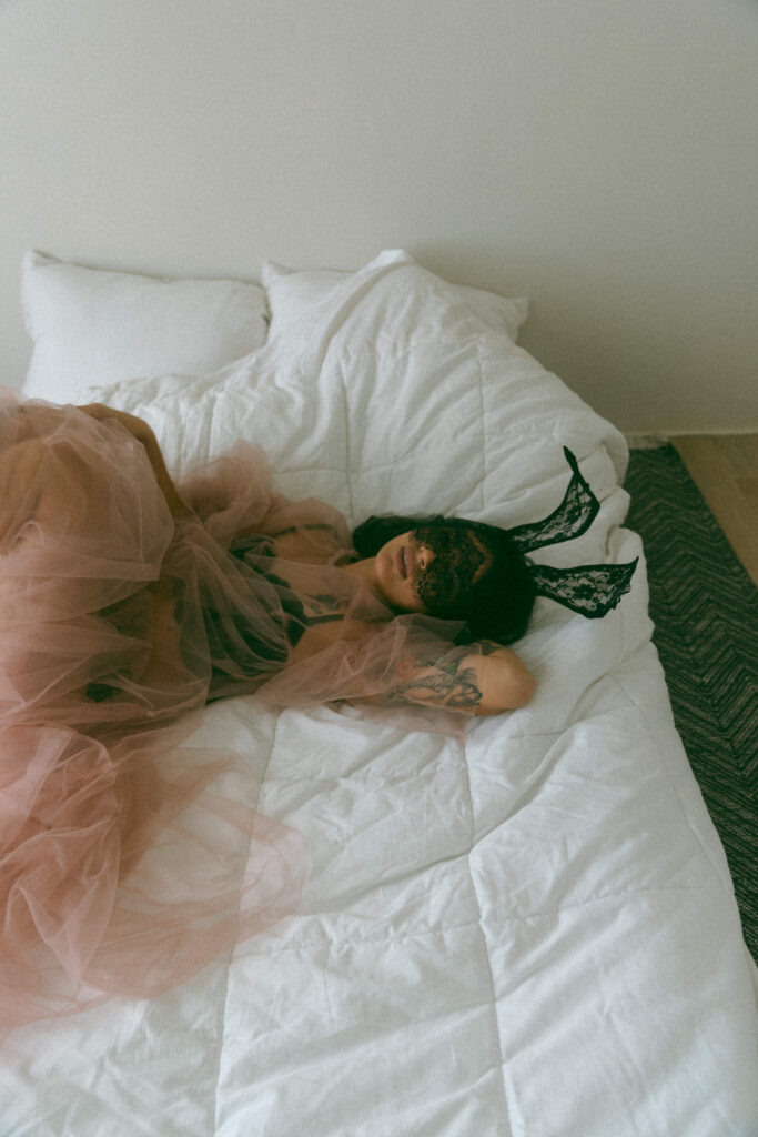 a woman laying in bed with a lace mask and ears and tulle shear dress at her boudoir session 