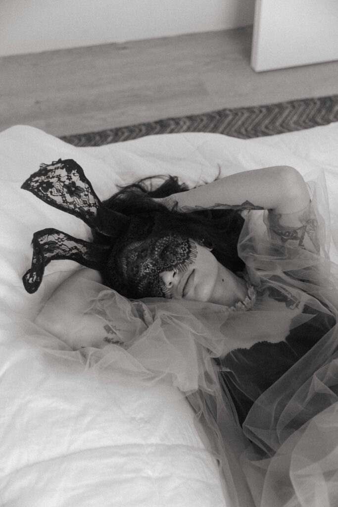 black and white of a woman laying in bed with a lace mask and ears and tulle shear dress at her boudoir session 