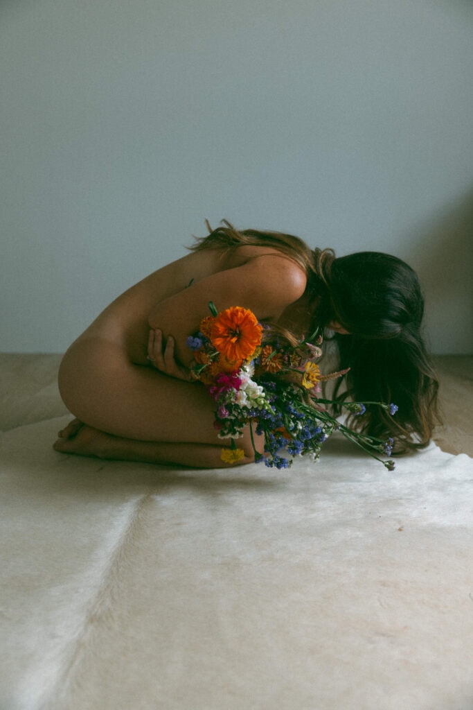 a naked woman curled over bright flowers at her boudoir session 