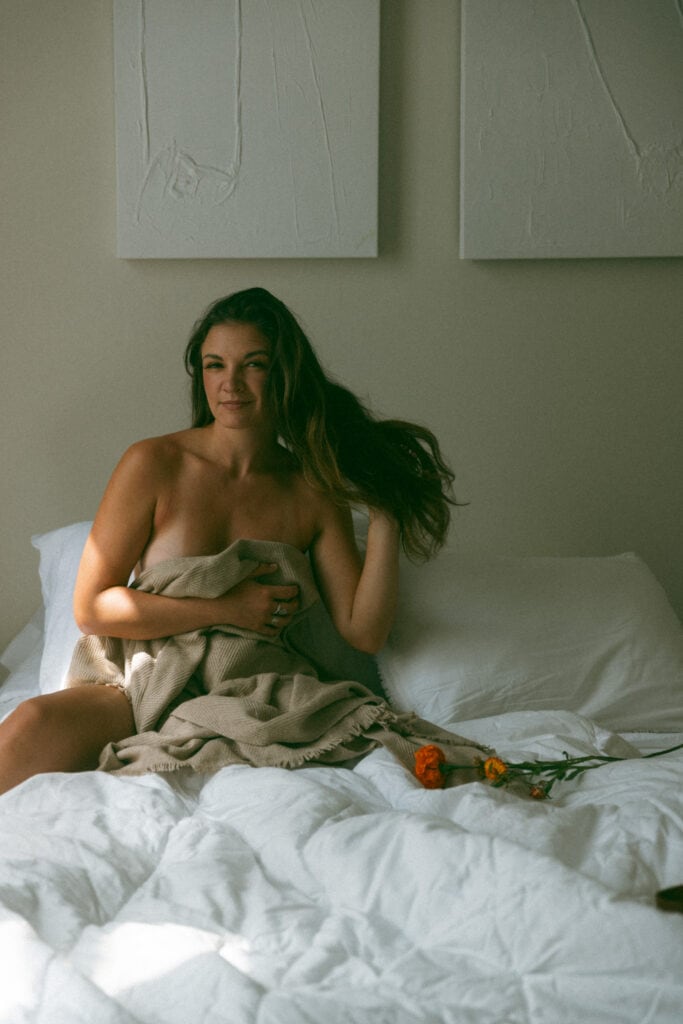 a naked woman in bed covering up with a blanket at her flower boudoir session 