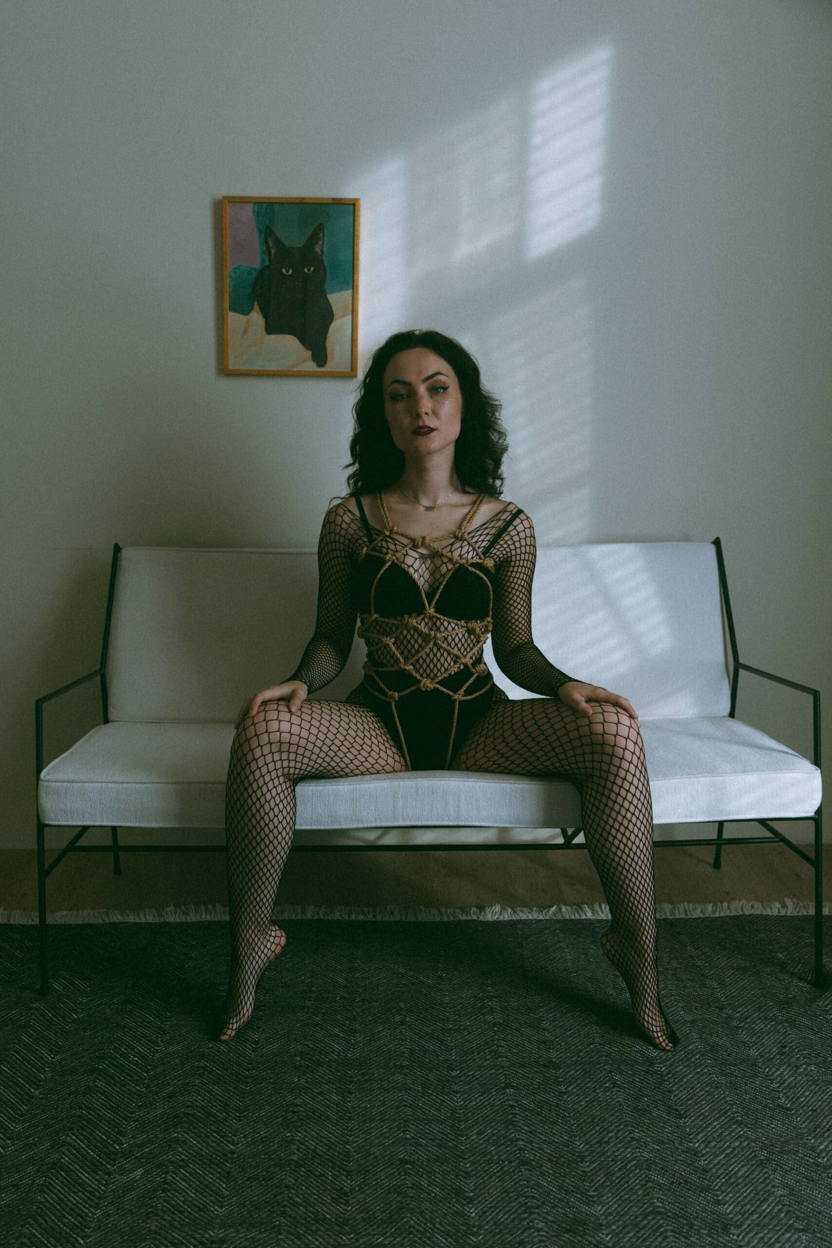 a woman in a fishnet bodysuit sitting on a couch with sensual shibari ropes tied around her whole torso