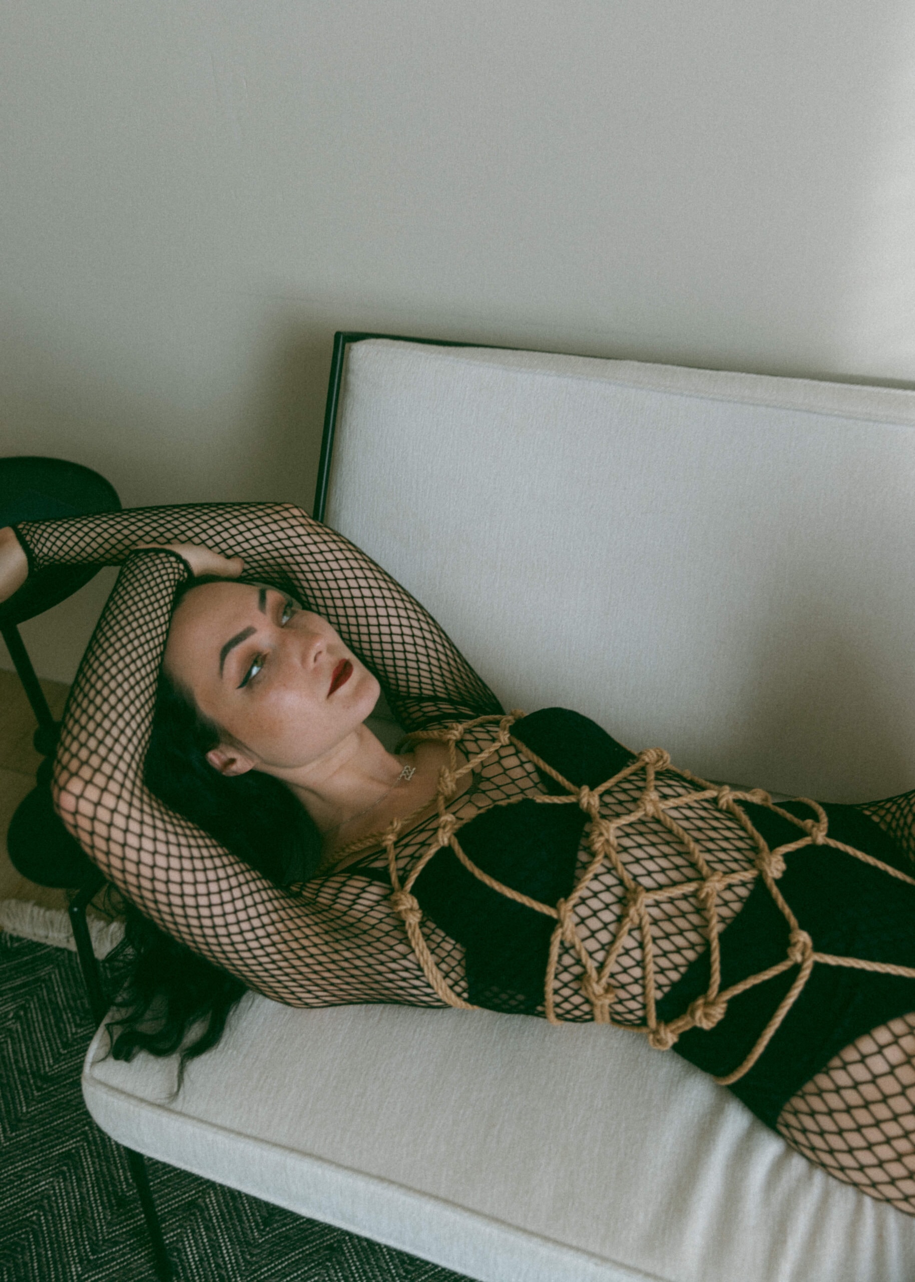 a woman in a fishnet bodysuit laying on a couch with shibari ropes tied around her whole torso