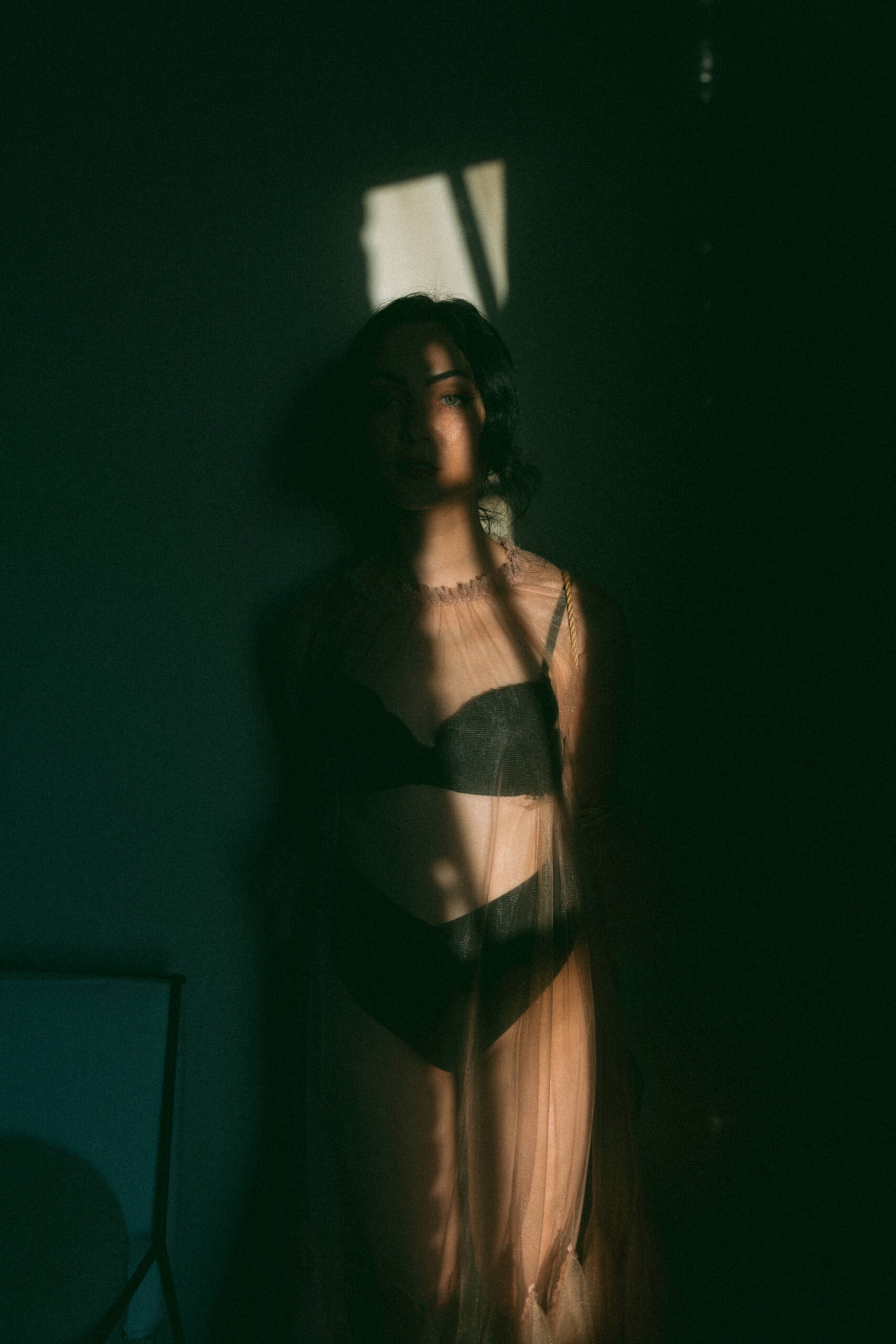 a woman sitting in the shadows with light streaking onto her body during a sensual shibari session