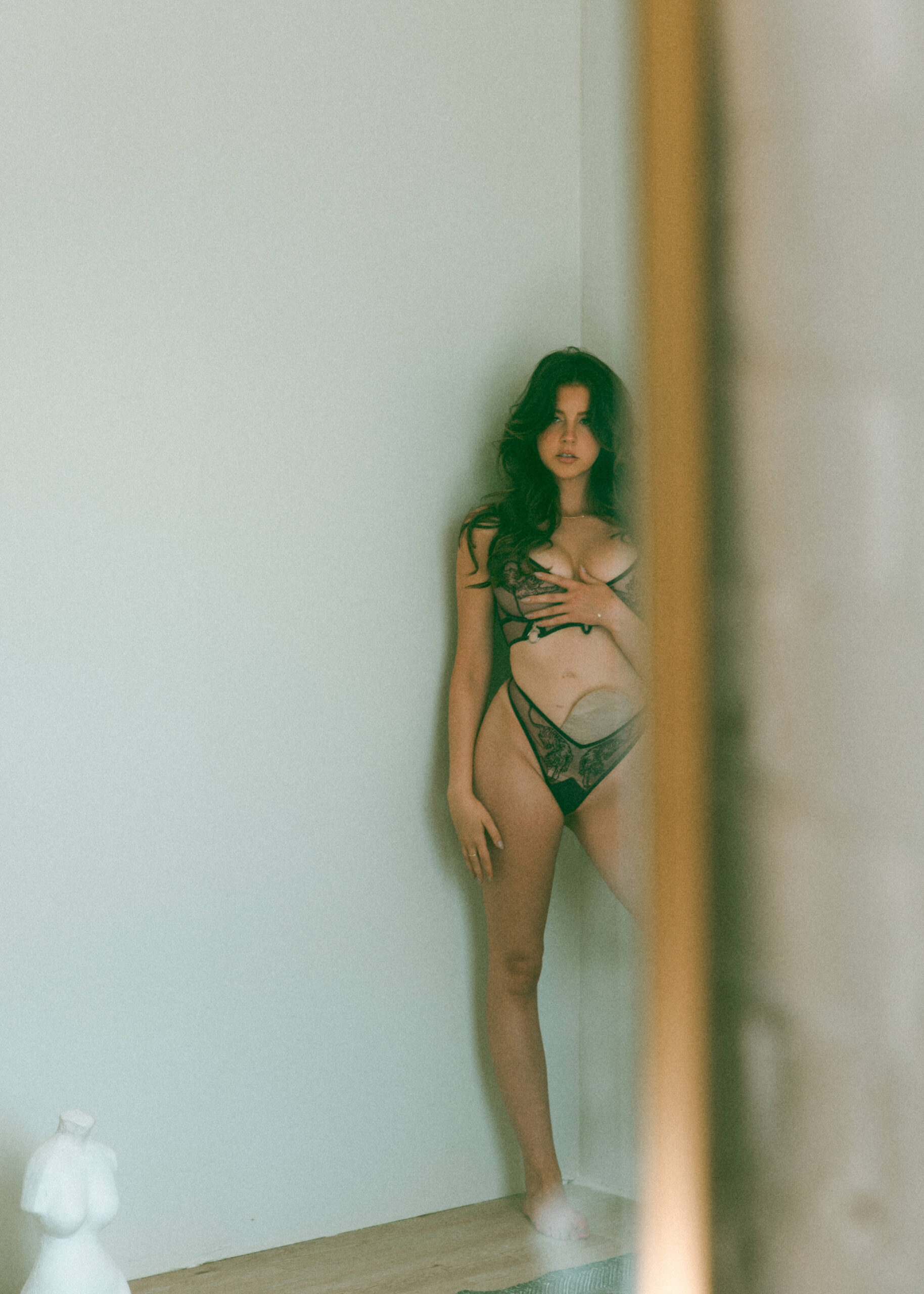 a woman with a medical device attached to her stomach, standing in the corner, wearing intimates and looking away from the camera during a boudoir session 