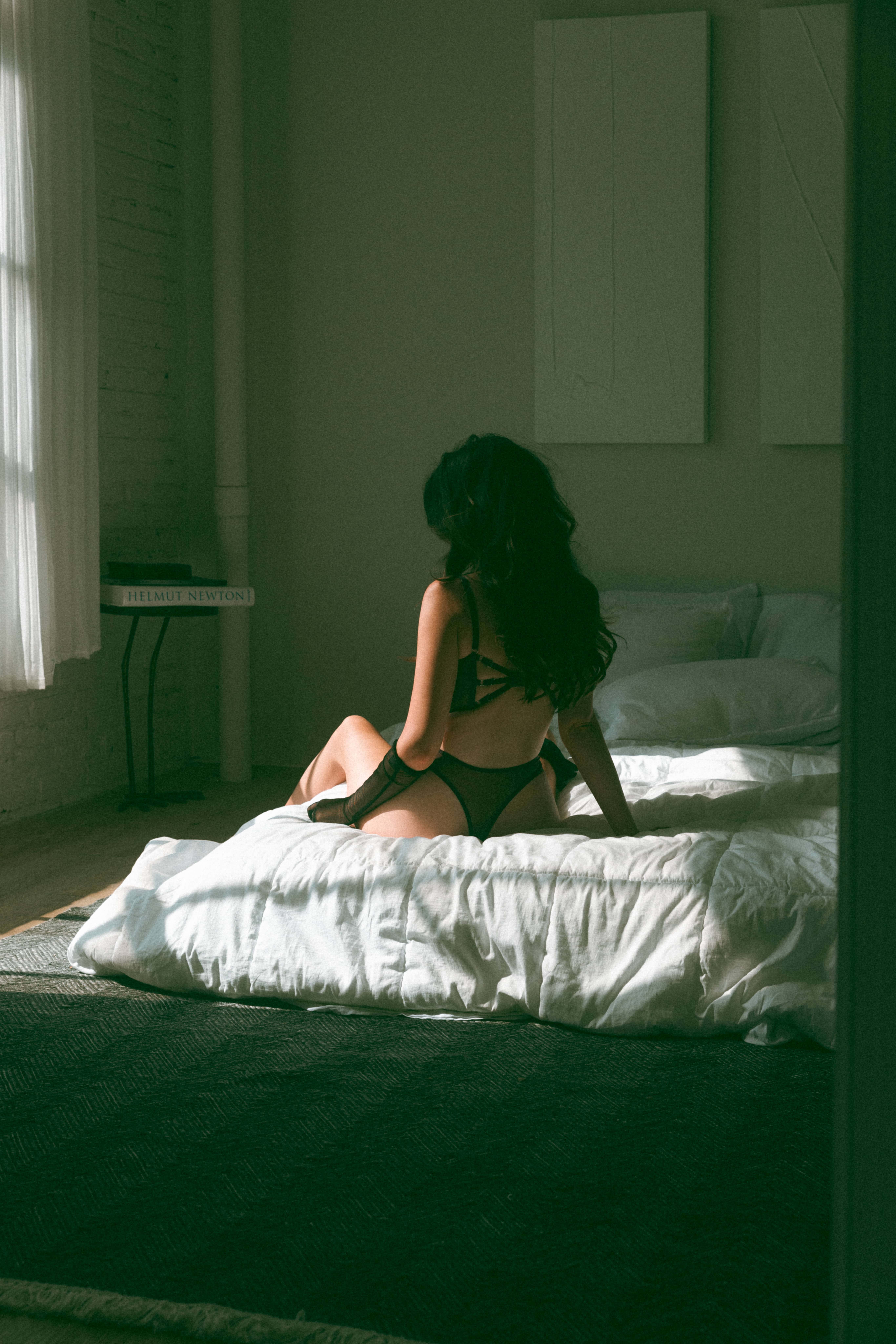 a woman sitting on a bed with her back to the camera, wearing intimates while the light from the windows shines on her