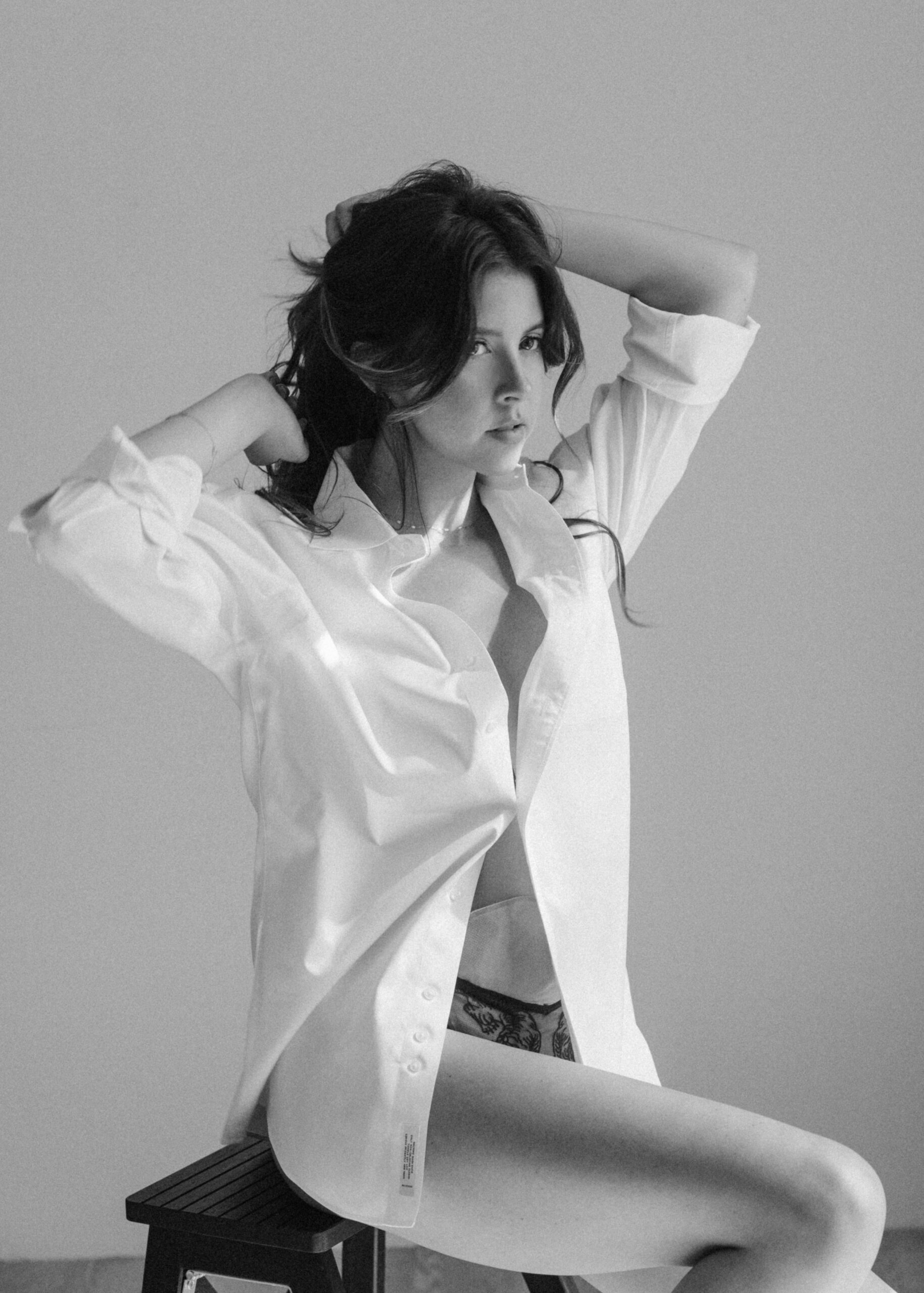 a woman sitting on a chair in an oversized white shirt and holding her hair during a boudoir session 