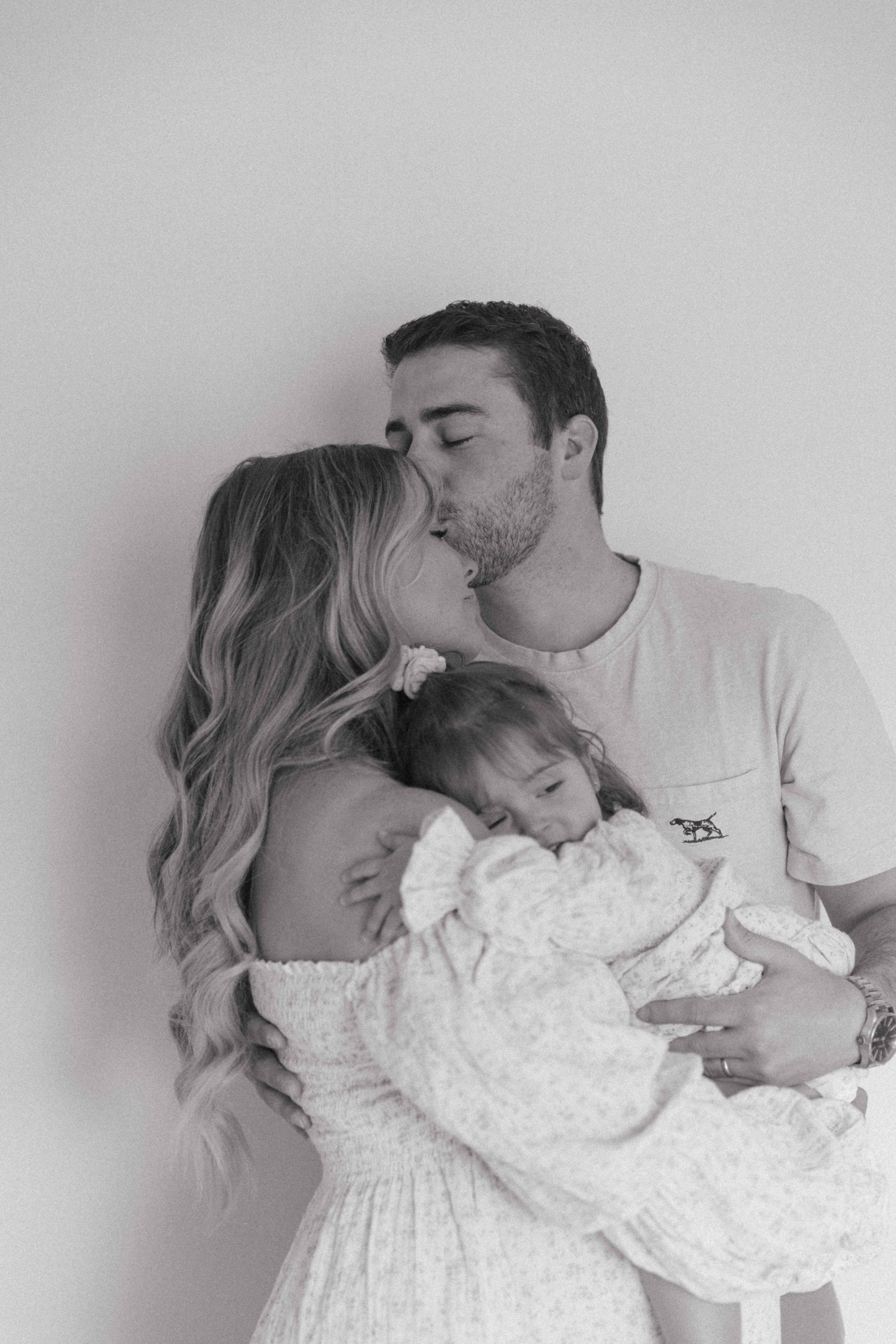 husband kissing wife's forehead while she holds their toddler during a studio maternity photoshoot