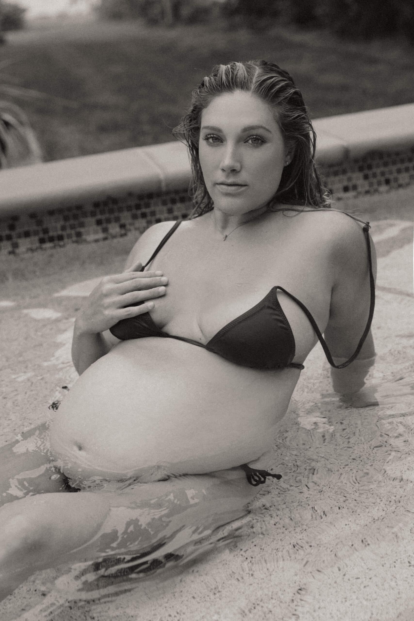 a pregnant woman sitting in the shallow part of the pool during an outdoor boudoir session