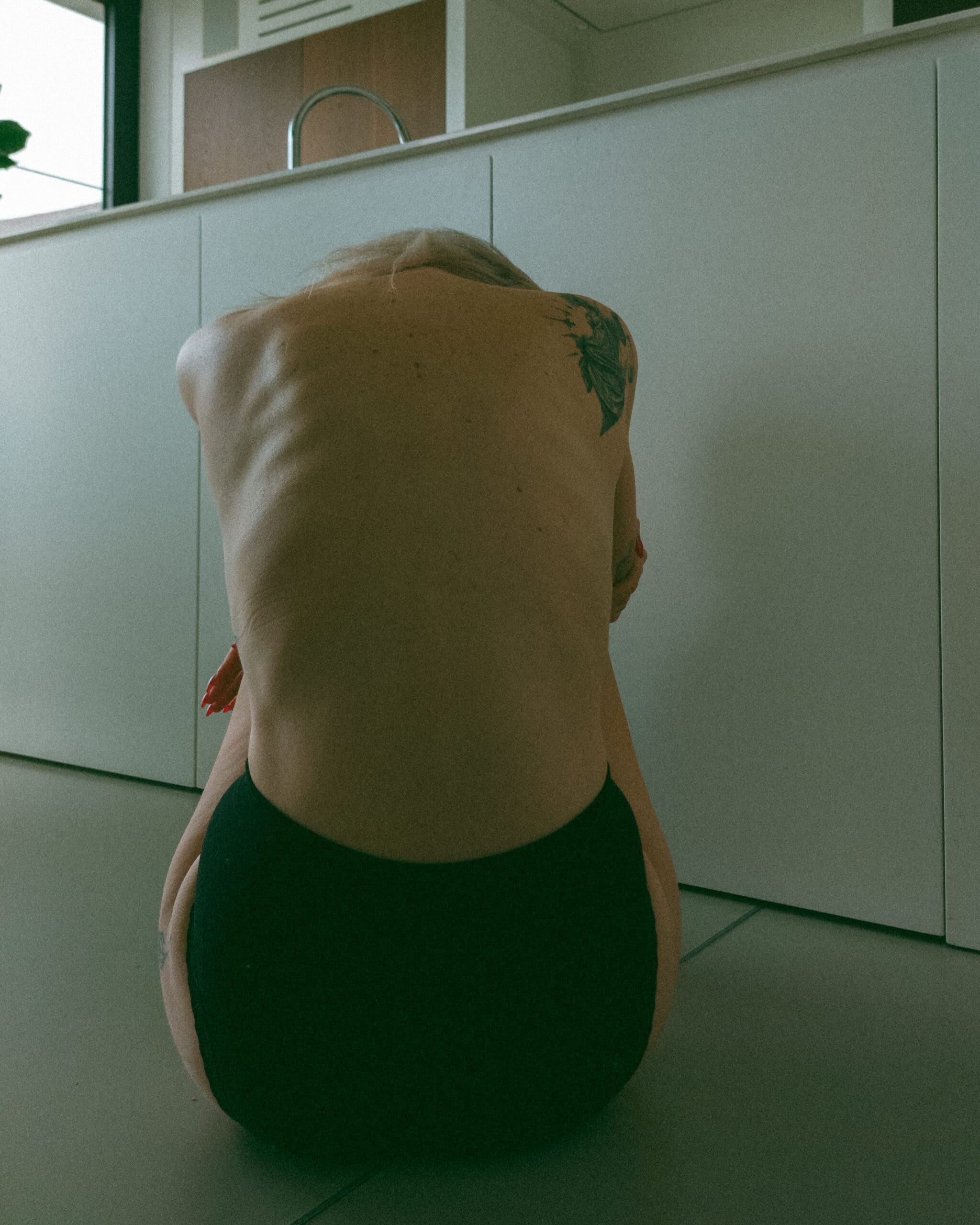 the back of a topless woman with her arms wrapped around her legs during an intimate portrait photography session at her home