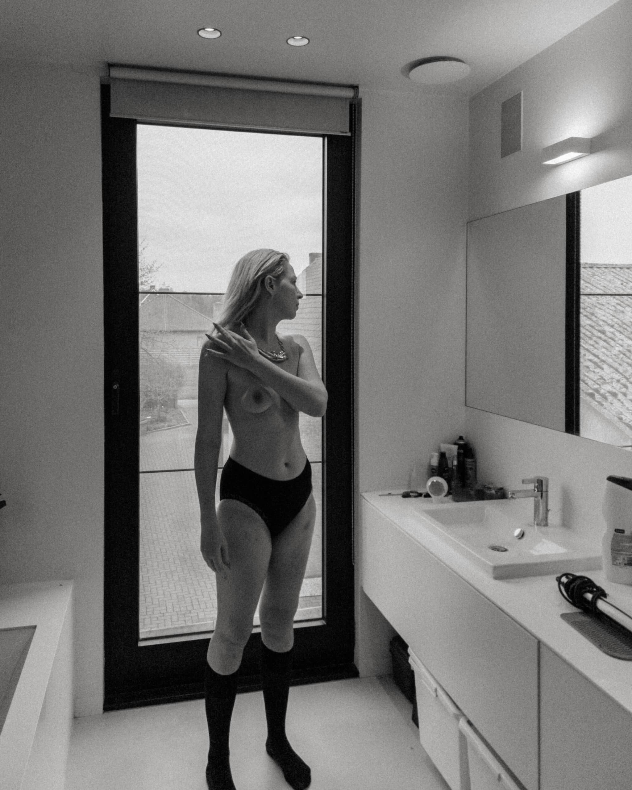 a topless woman standing in her bathroom brushing her hair off her shoulder during an intimate portrait photography session at her home