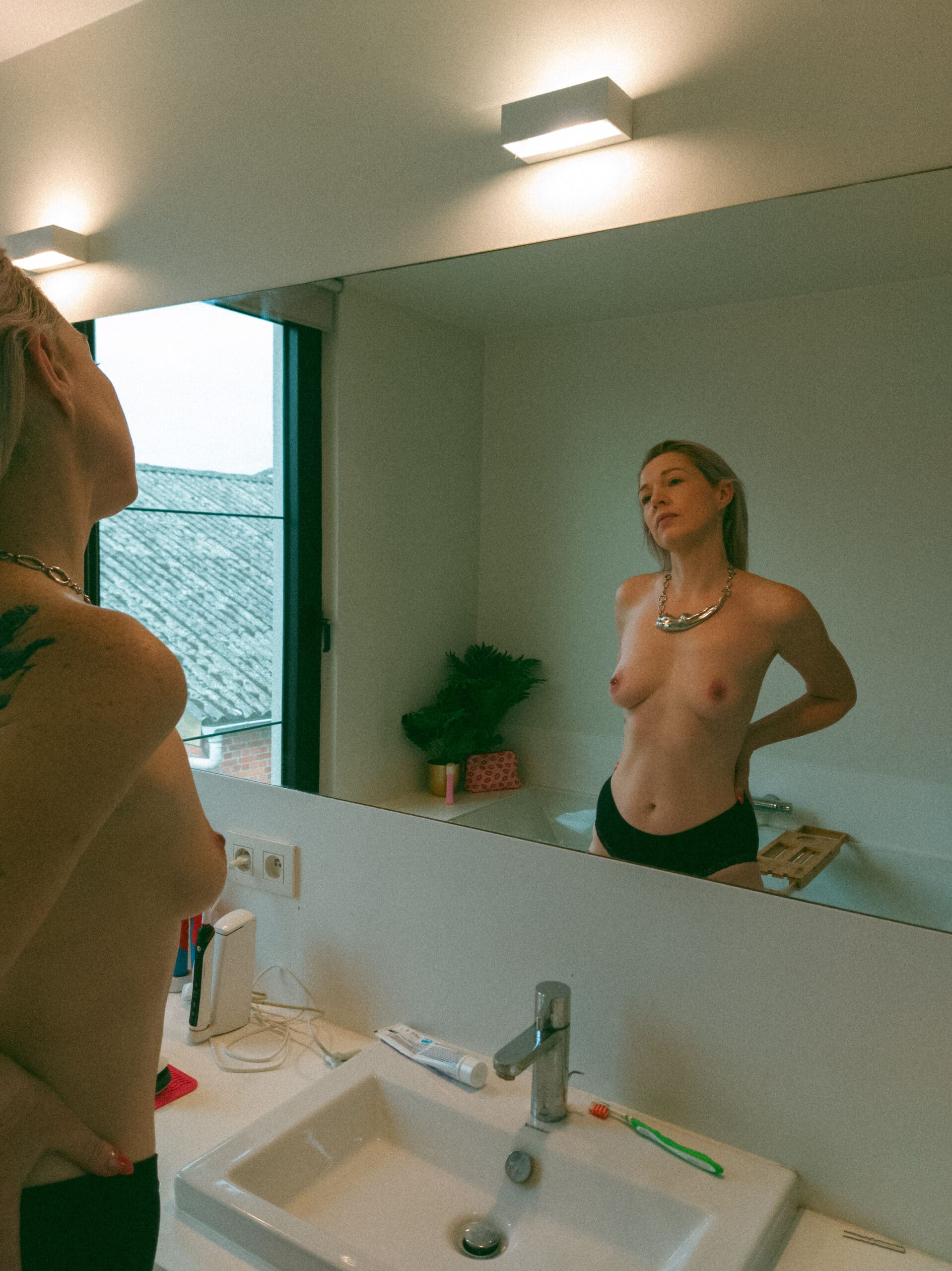 a topless woman with her hands on her waist looking into the mirror during an intimate portrait photography session at her home