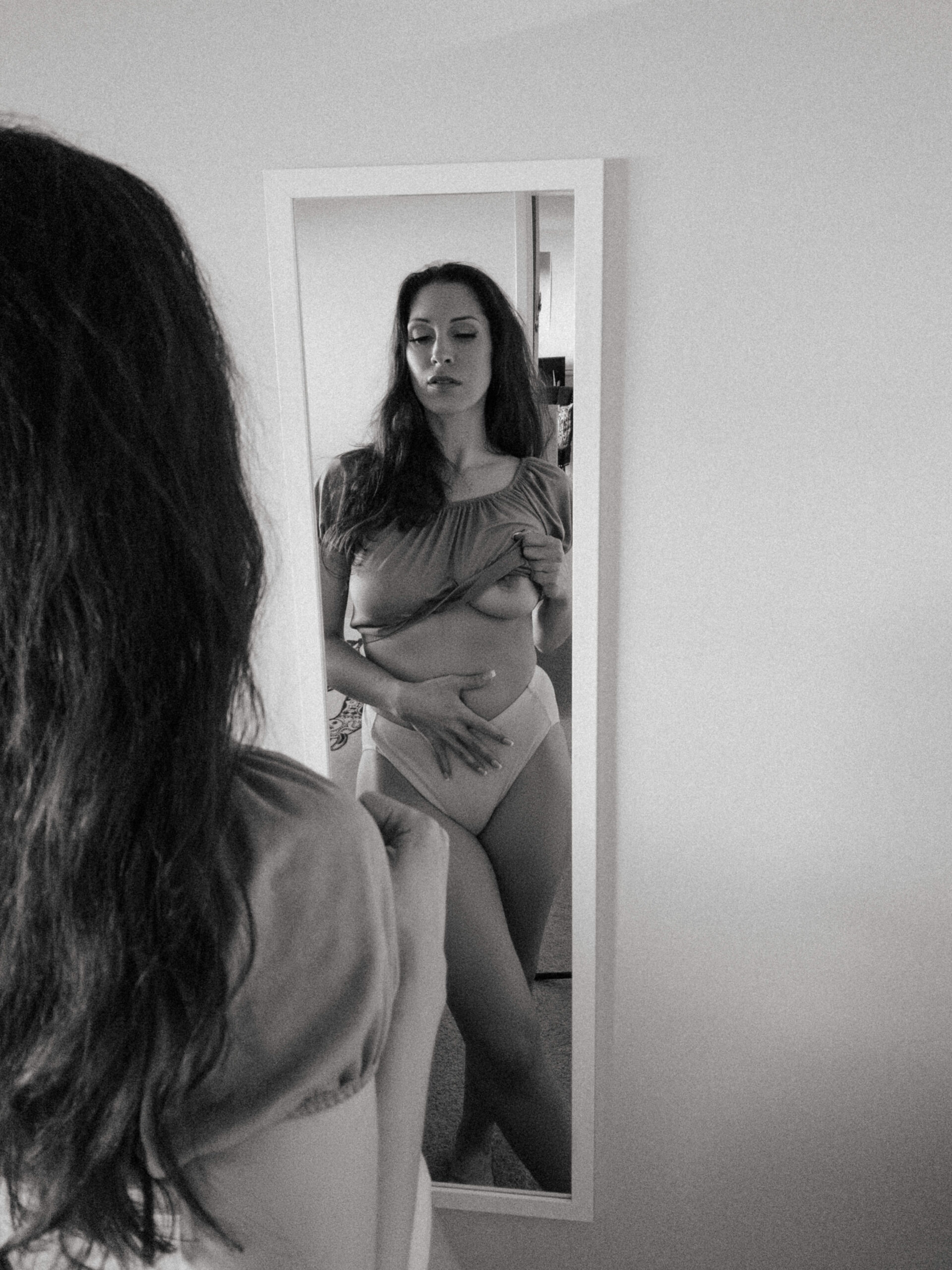 a woman holding her shirt up while looking into the mirror during at home boudoir photos 