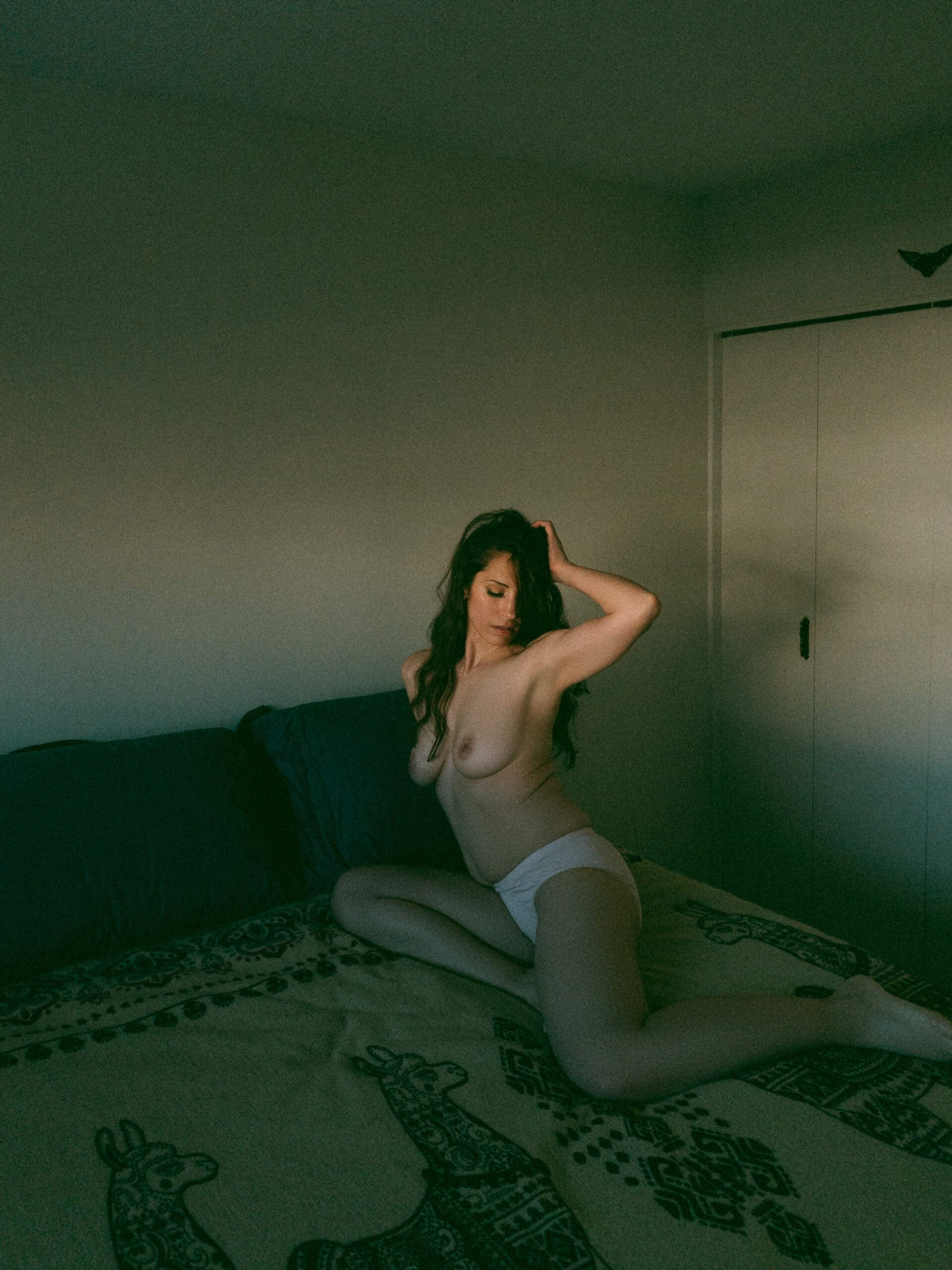 a topless woman with her hand on her head while sitting in her bed during at home boudoir photos 