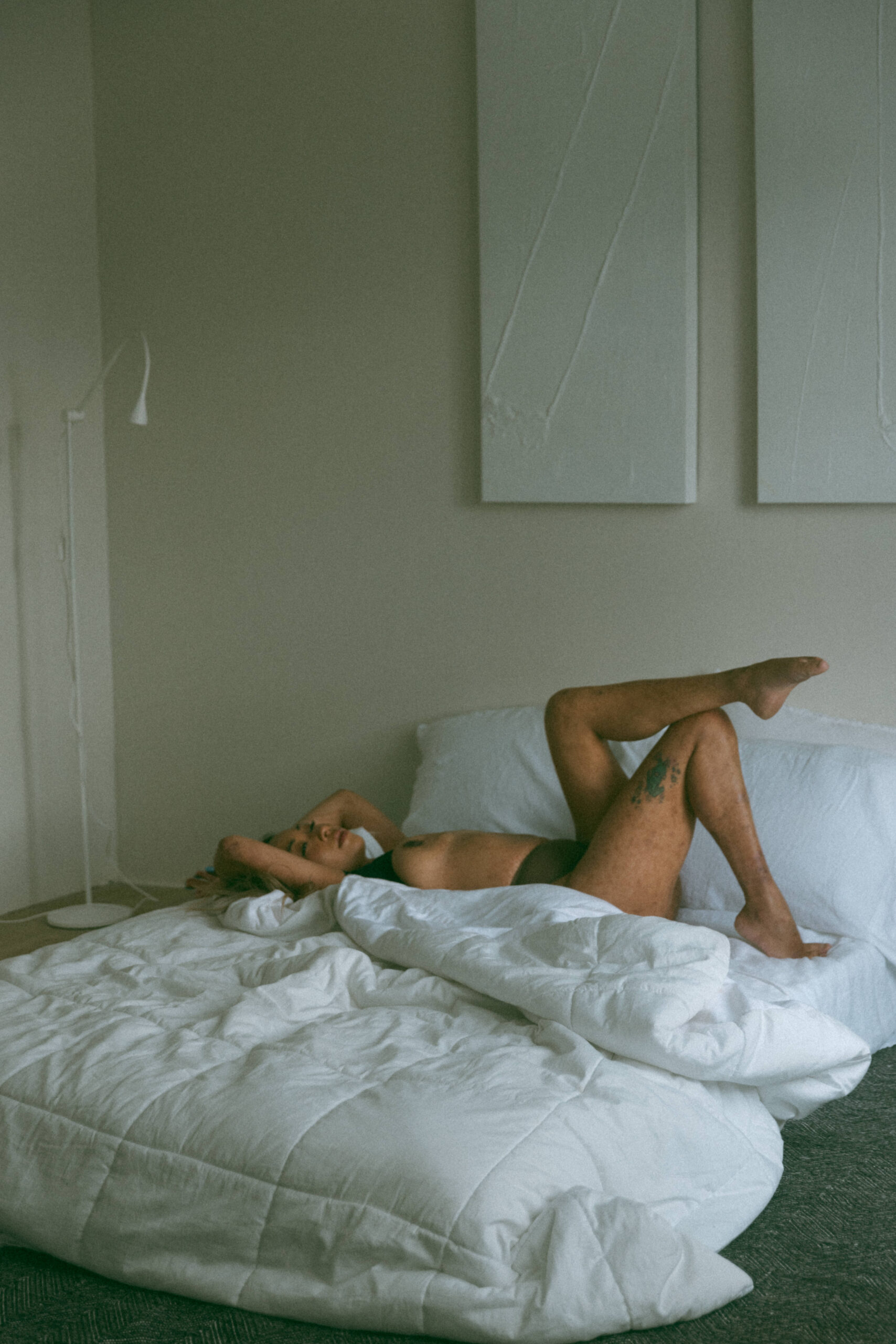 a topless woman laying in a bed crossing her legs during an artistic collaboration photoshoot 
